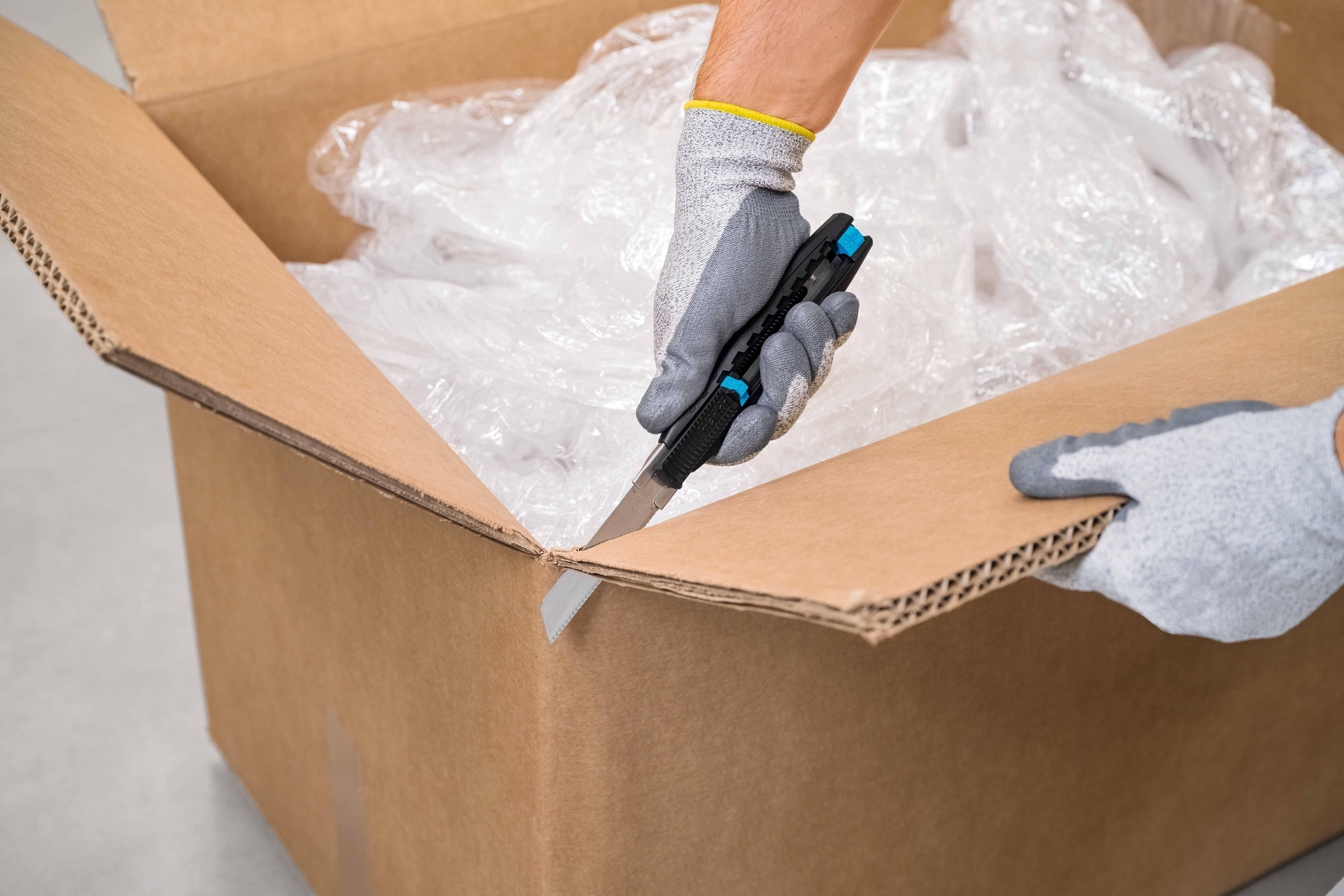 Une personne ouvre un carton à l'aide d'un cutter, à l'intérieur se trouvent des emballages en plastique.