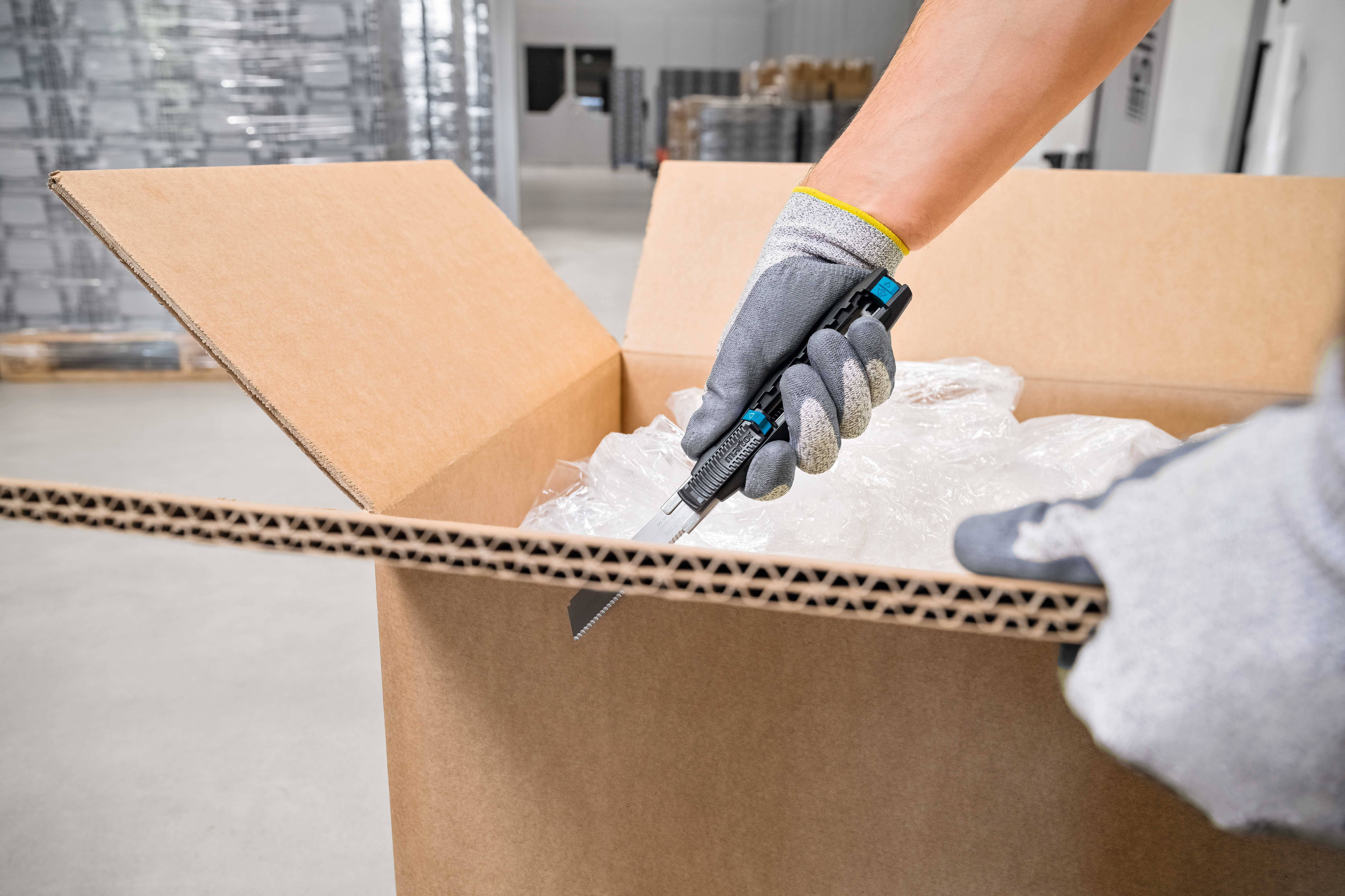 Une personne portant des gants de protection ouvre un carton à l'aide d'un couteau utilitaire. Des étagères industrielles sont visibles en arrière-plan.