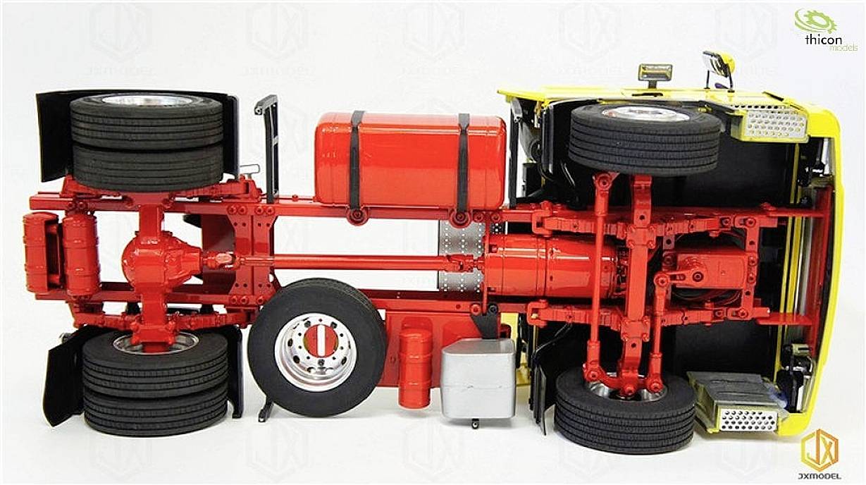 Un camion-modèle est couché sur le côté, montrant son châssis. On peut voir des essieux, des pneus, un réservoir de carburant rouge et des pièces de moteur.