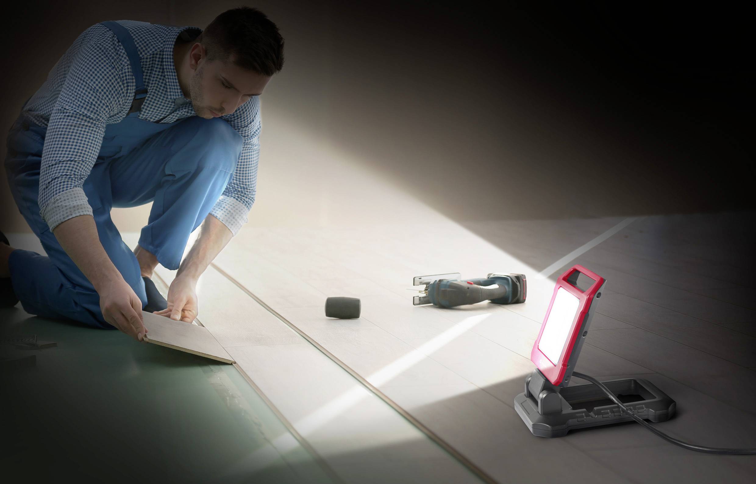 Un homme pose un revêtement de sol stratifié dans une pièce, éclairé par une lampe de travail. Des outils sont posés à côté de lui sur le sol.