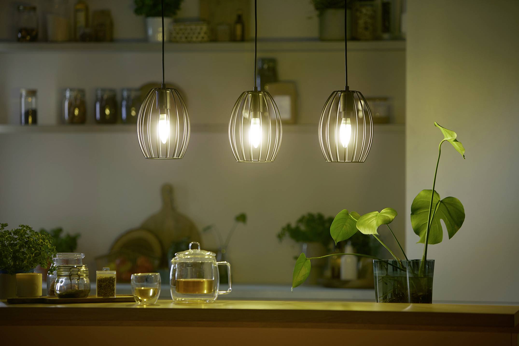 Trois lampes suspendues éclairent une ligne de cuisine avec une théière, une tasse et une plante. En arrière-plan, des étagères contenant des bocaux d'épices sont visibles.