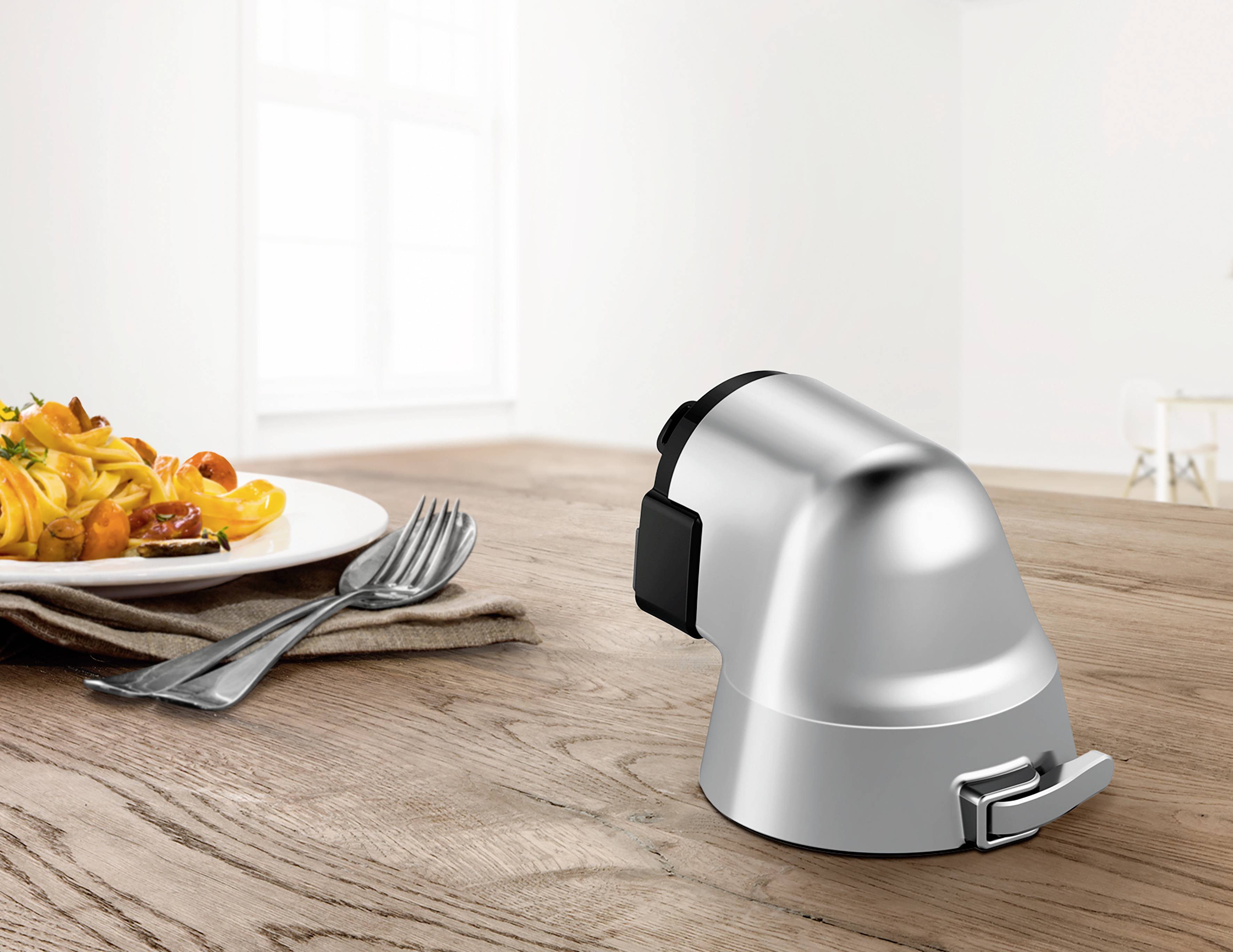 Un coupe-ail argenté est posé sur une table en bois à côté d'une assiette de pâtes et de couverts.