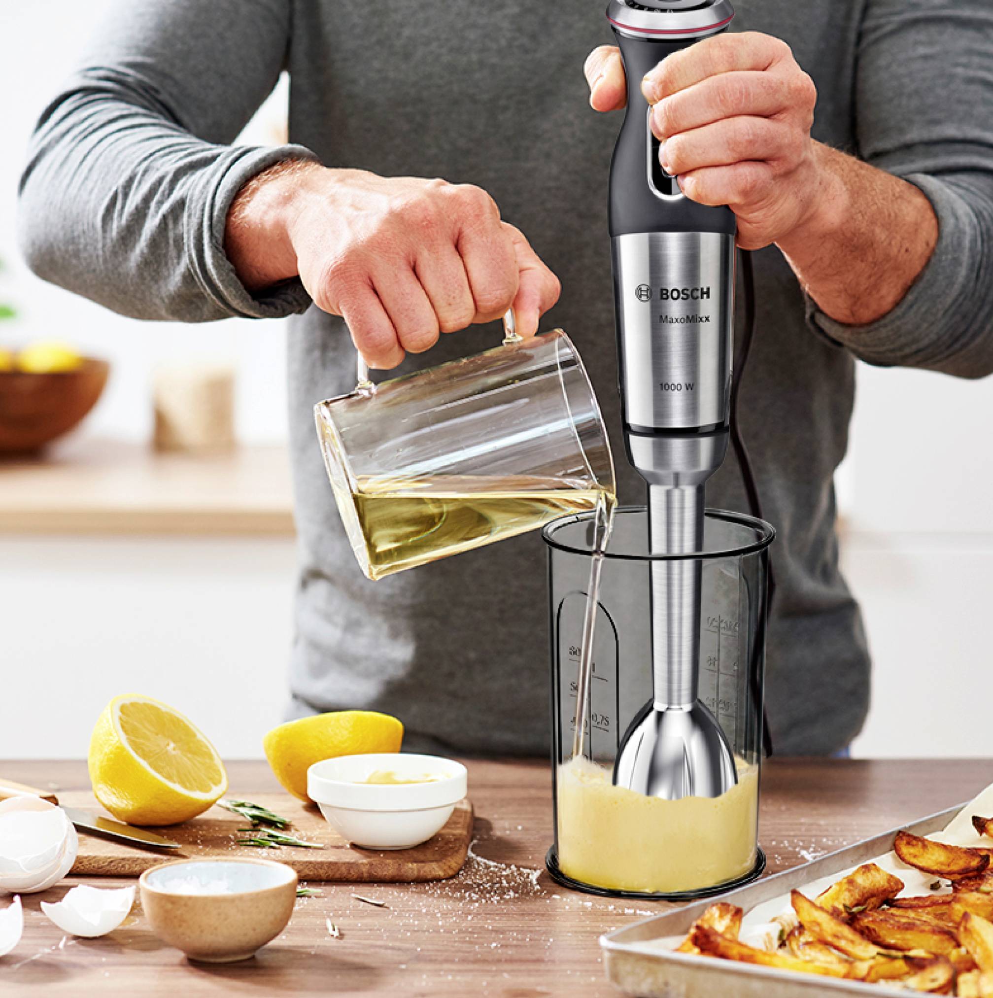 Une personne utilise un mixeur plongeant pour réduire un liquide en purée dans un récipient. À côté se trouvent des citrons, des œufs et un moule à gâteau.