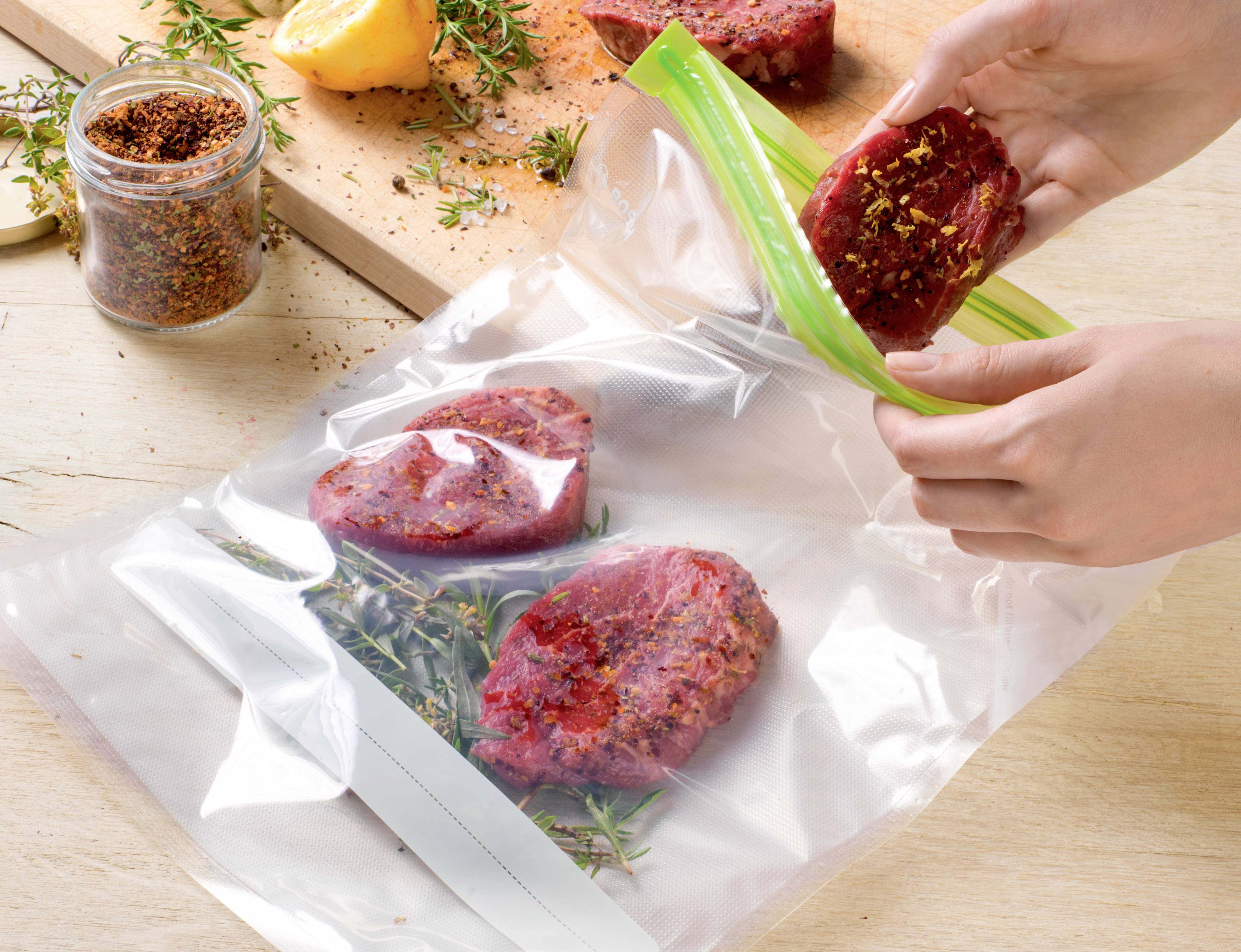 Une personne place des steaks assaisonnés dans un sac sous vide. Des épices et des herbes sont visibles en arrière-plan.