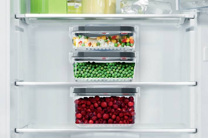 Réfrigérateur ouvert contenant trois récipients transparents remplis d'aliments surgelés : des légumes en haut, des petits pois au milieu, des baies rouges en bas.