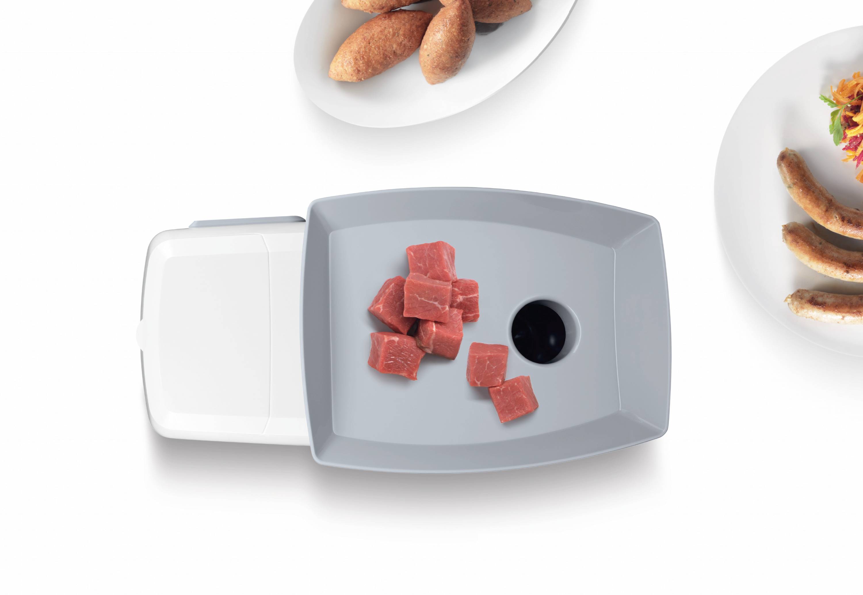 Assiette grise avec des dés de viande crus, avec en arrière-plan une assiette de pain et de saucisse.