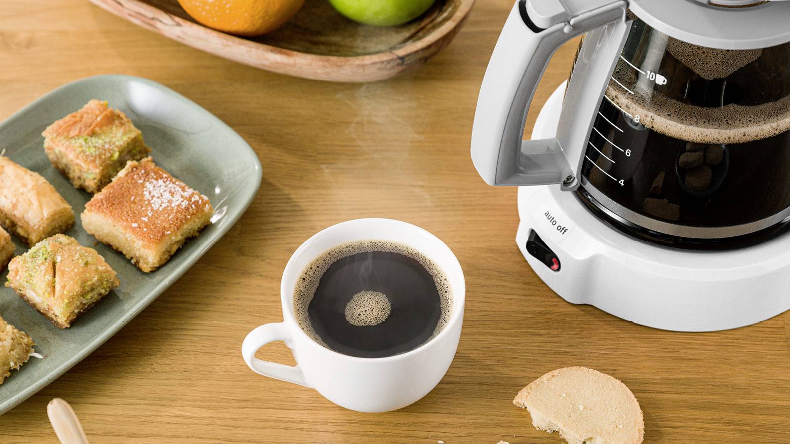 Table du petit-déjeuner avec une tasse de café, des parts de gâteau et une machine à café, entourée d'un plateau de fruits avec des oranges et des pommes.
