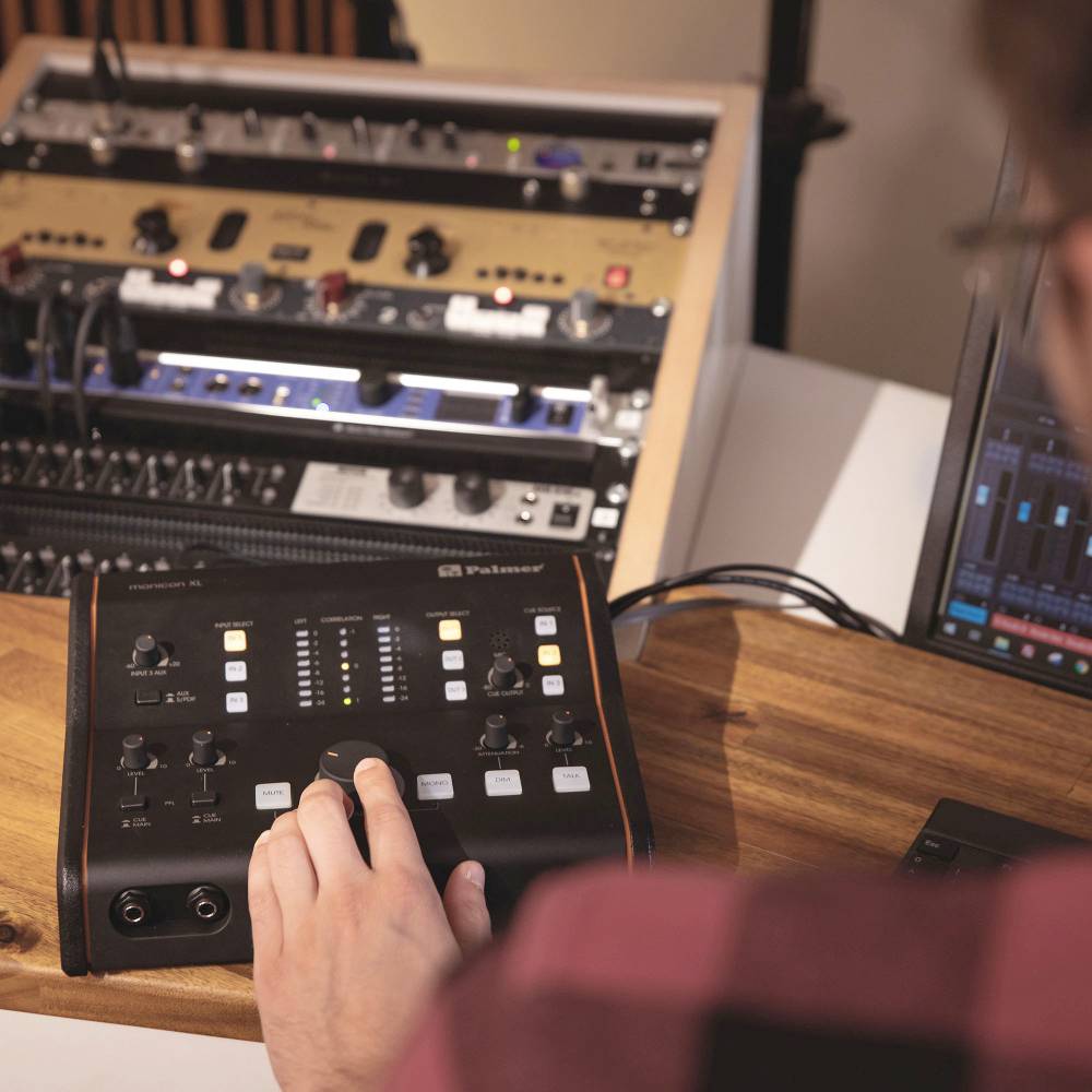 Une personne manipule une console de mixage audio dans un studio, entourée de divers équipements audio et d'un écran d'ordinateur.