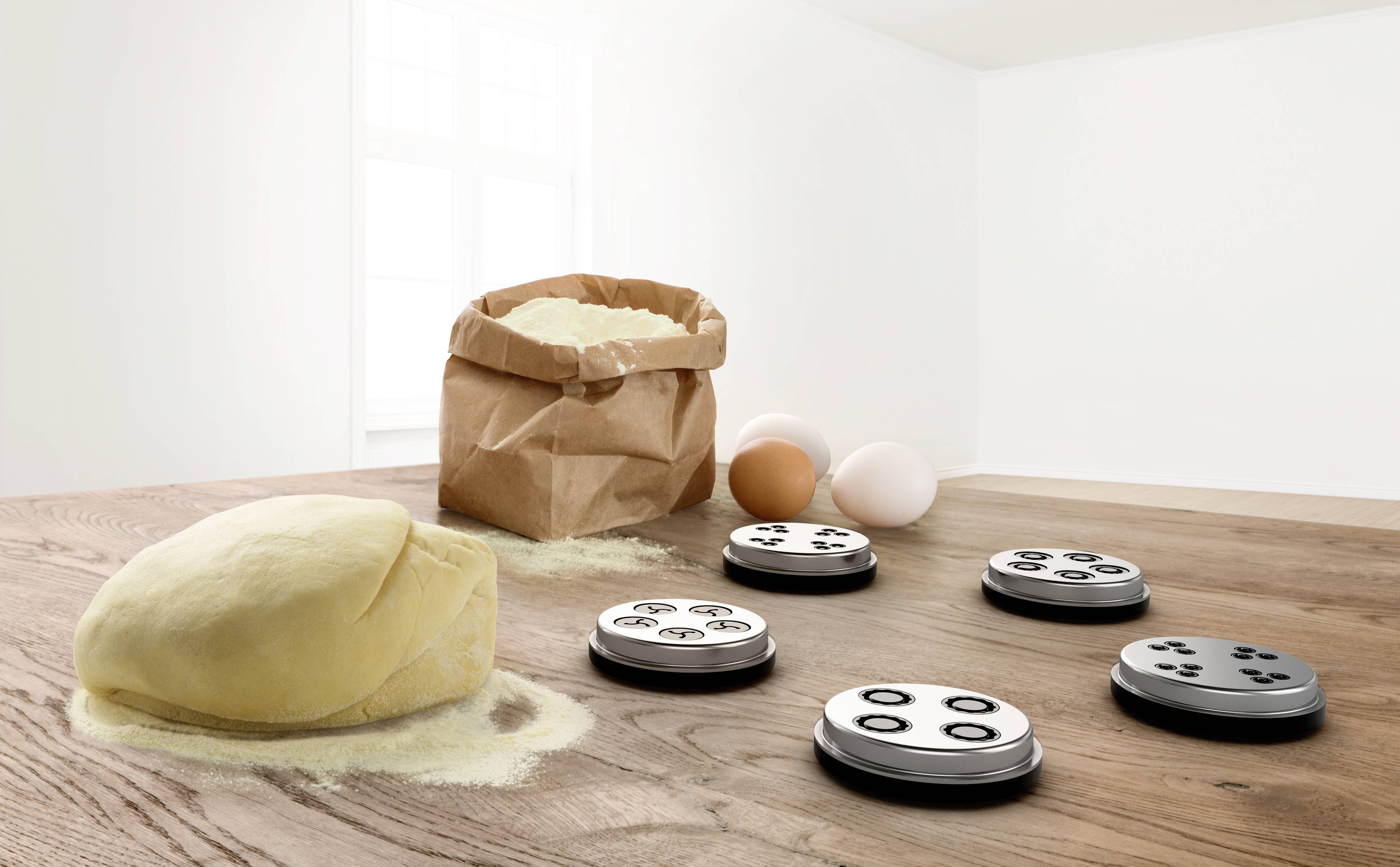 Une boule de pâte, cinq disques métalliques aux motifs différents, un sac de farine ouvert et deux œufs sur une table en bois.