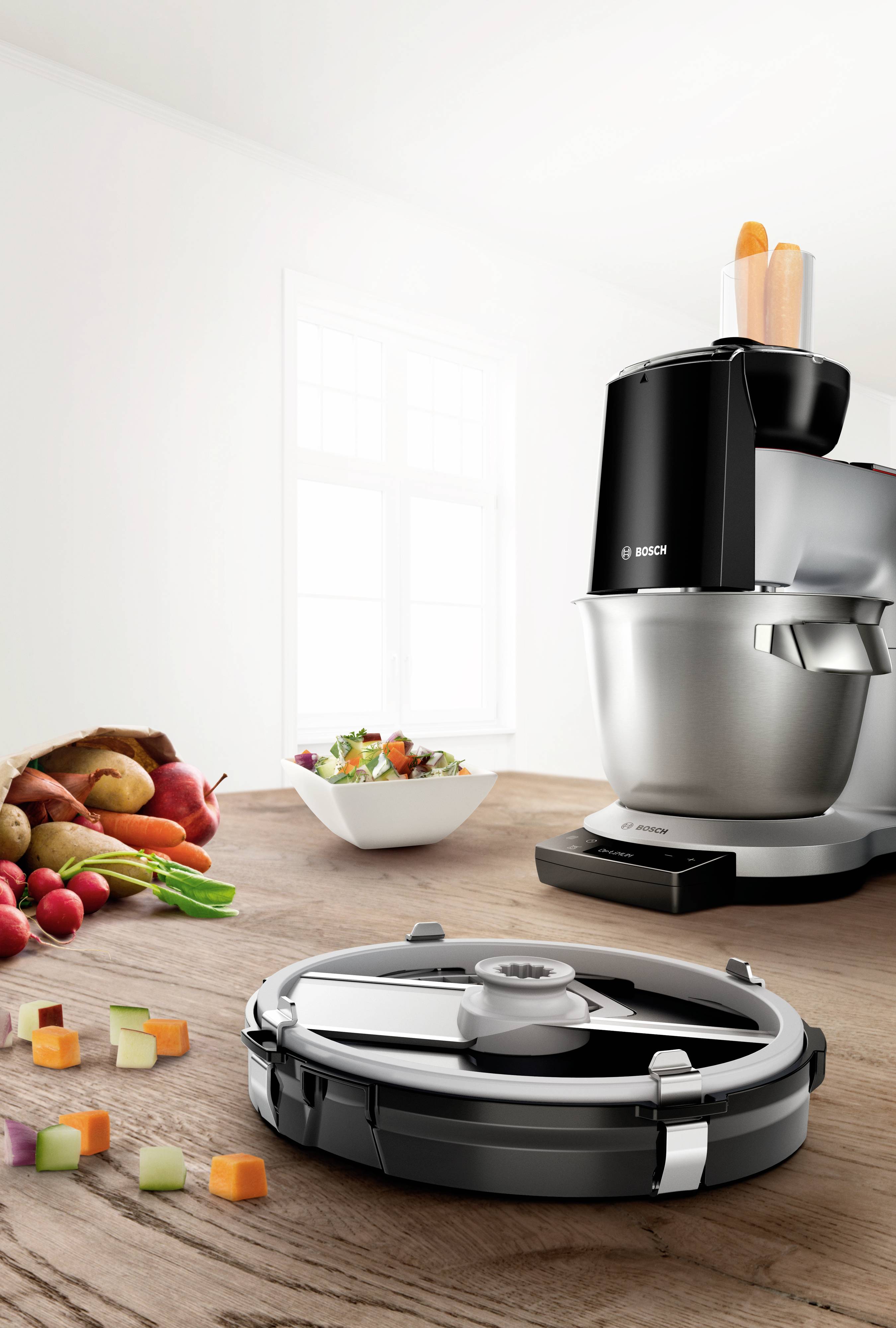 Un robot de cuisine noir sur une table en bois, avec des morceaux de légumes, un bol de salade et des légumes frais en arrière-plan.