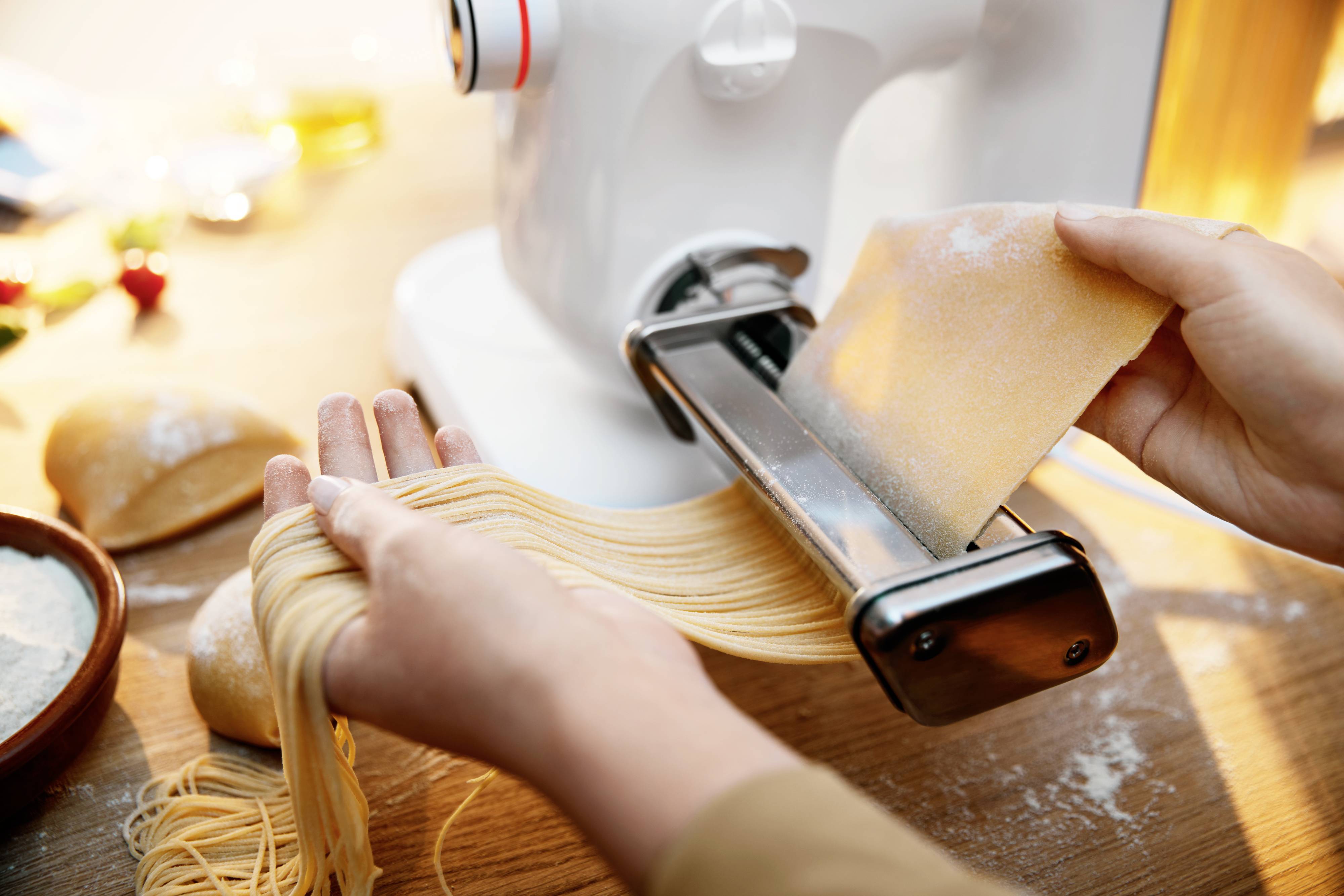 Des mains façonnent la pâte à pâtes avec une machine à pâtes. Des pâtes fraîches sont découpées, saupoudrées de farine sur une planche en bois.