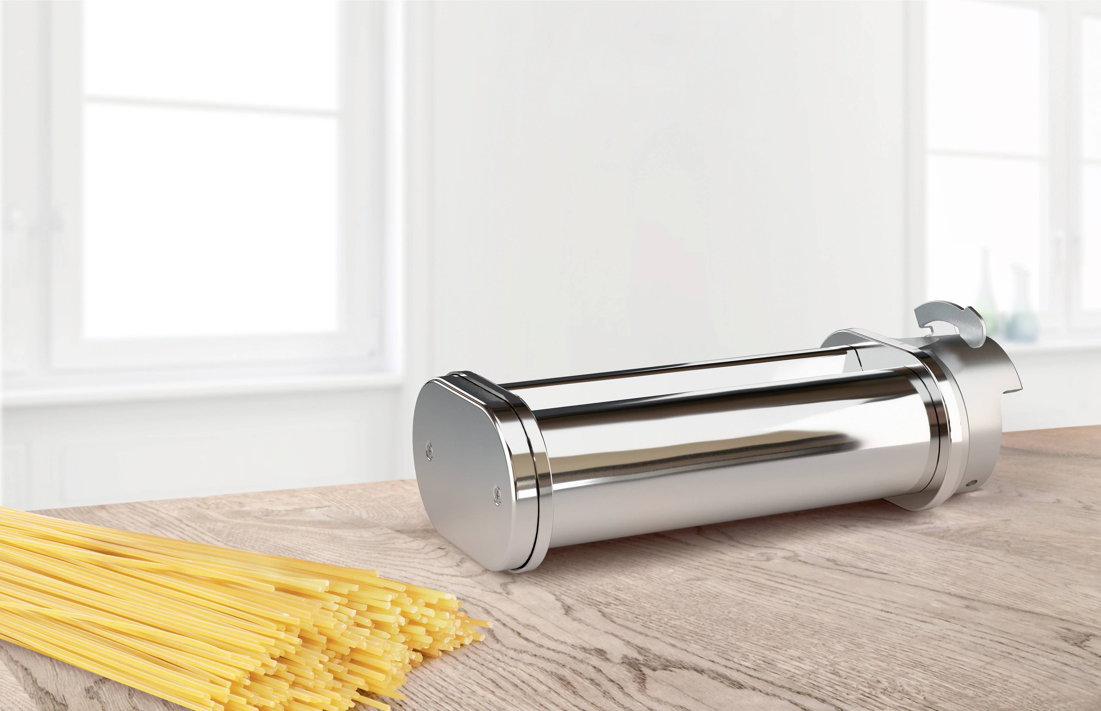 Un embout-poussoir argenté est posé sur une table en bois à côté d'un paquet de spaghetti crus, avec une fenêtre en arrière-plan.