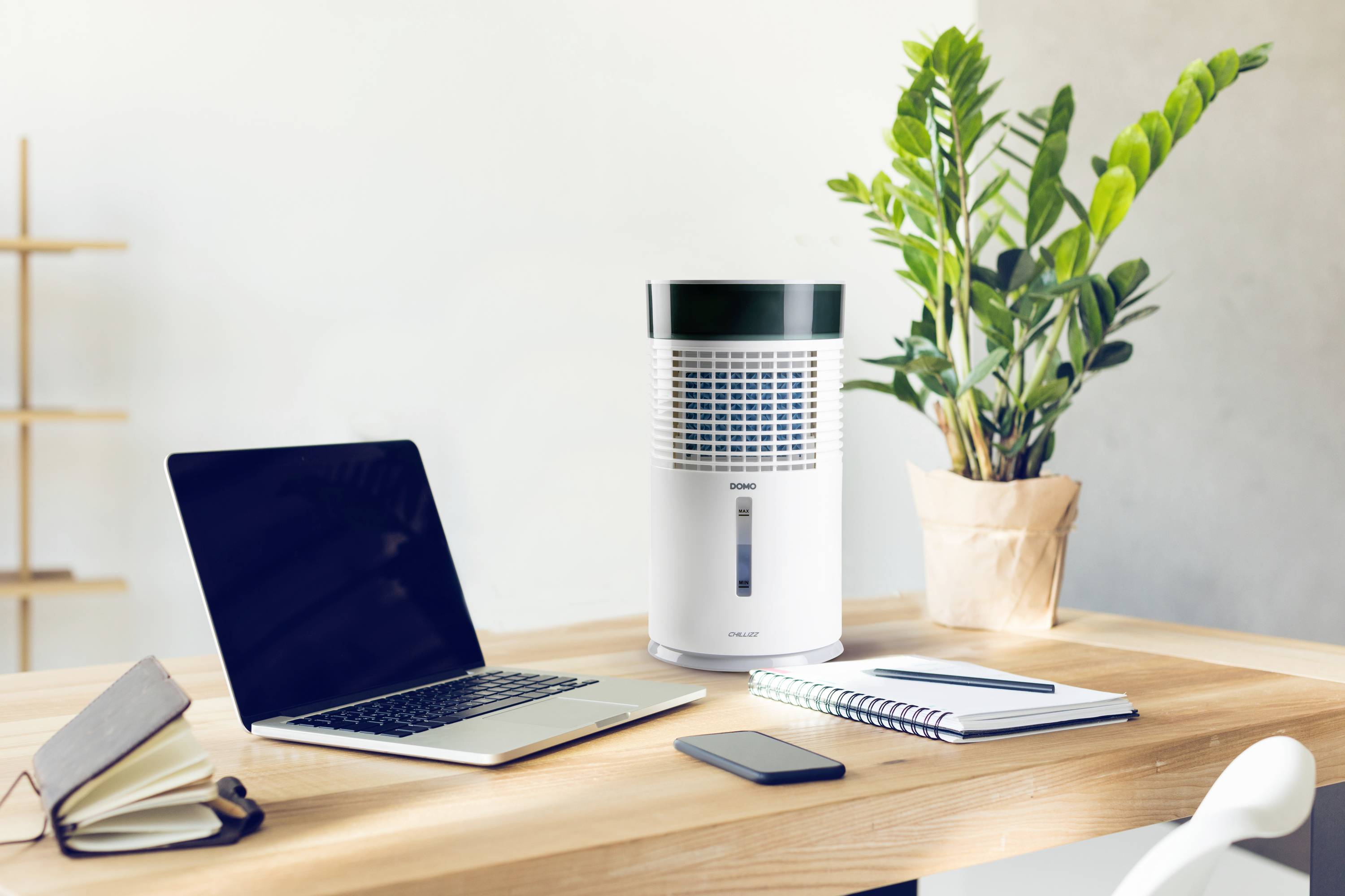 Un purificateur d'air est posé sur un bureau à côté d'un ordinateur portable, d'un carnet de notes et d'un smartphone. Une plante verte se trouve en arrière-plan.