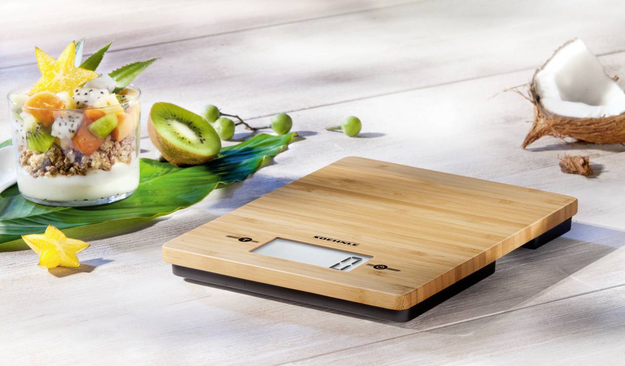 Balance de cuisine numérique en bois posée sur une table, avec des fruits frais et du yaourt en arrière-plan.