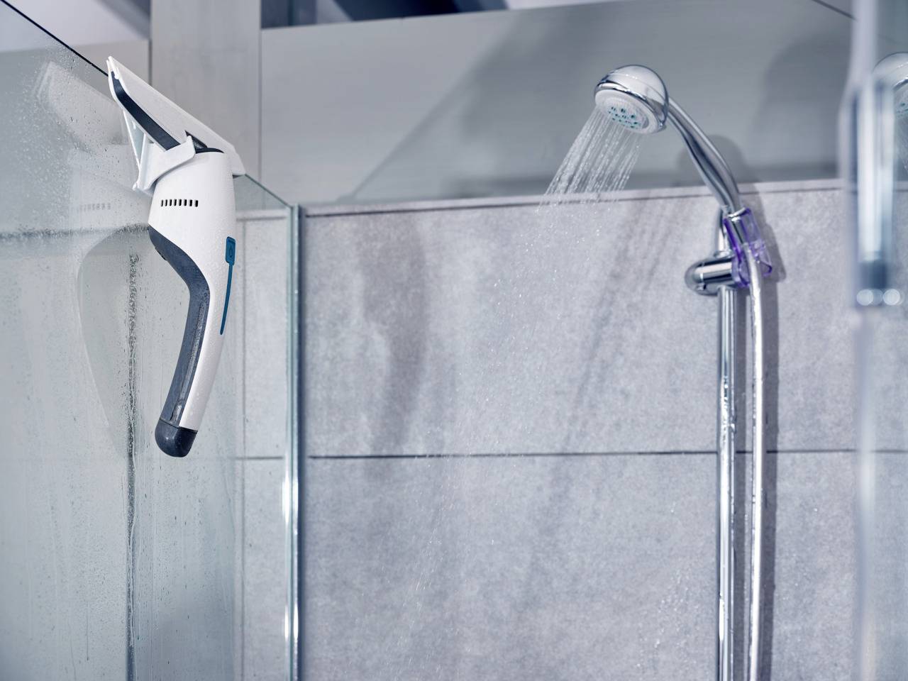 Un pommeau de douche projette de l'eau dans une douche moderne aux carreaux gris. Un rasoir est accroché au mur de la douche.