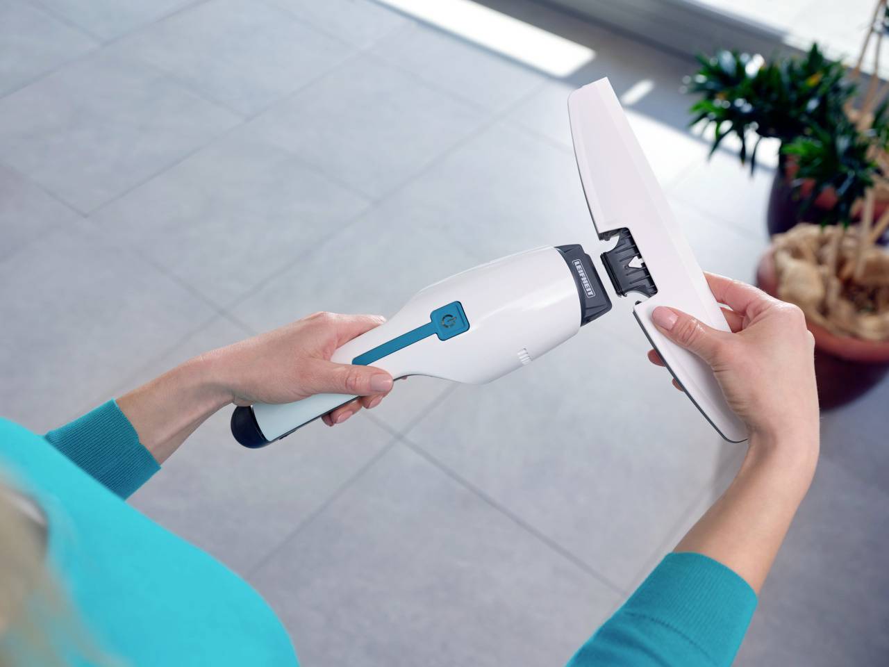 Une personne tient un aspirateur de fenêtre électrique au design blanc et bleu sur un sol carrelé.