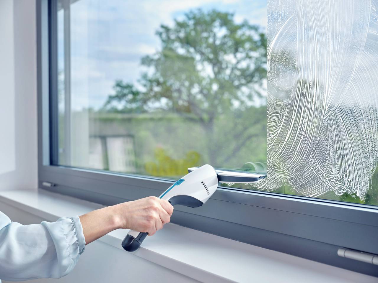 Une personne nettoie la fenêtre avec un produit de nettoyage à la main. En arrière-plan, on aperçoit des arbres et un ciel nuageux.