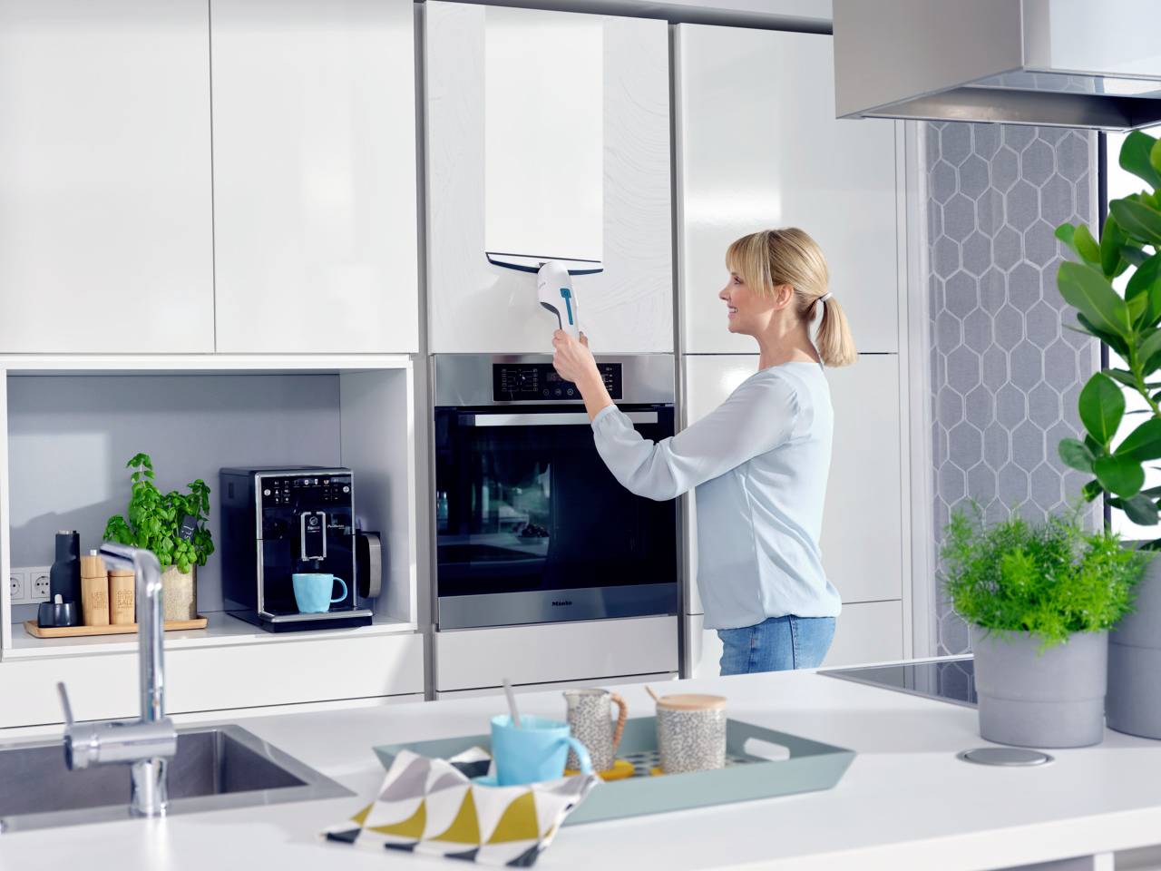 Une femme nettoie une porte de placard de cuisine avec un appareil de nettoyage électrique. À l'arrière-plan, on aperçoit une machine à café et un four.