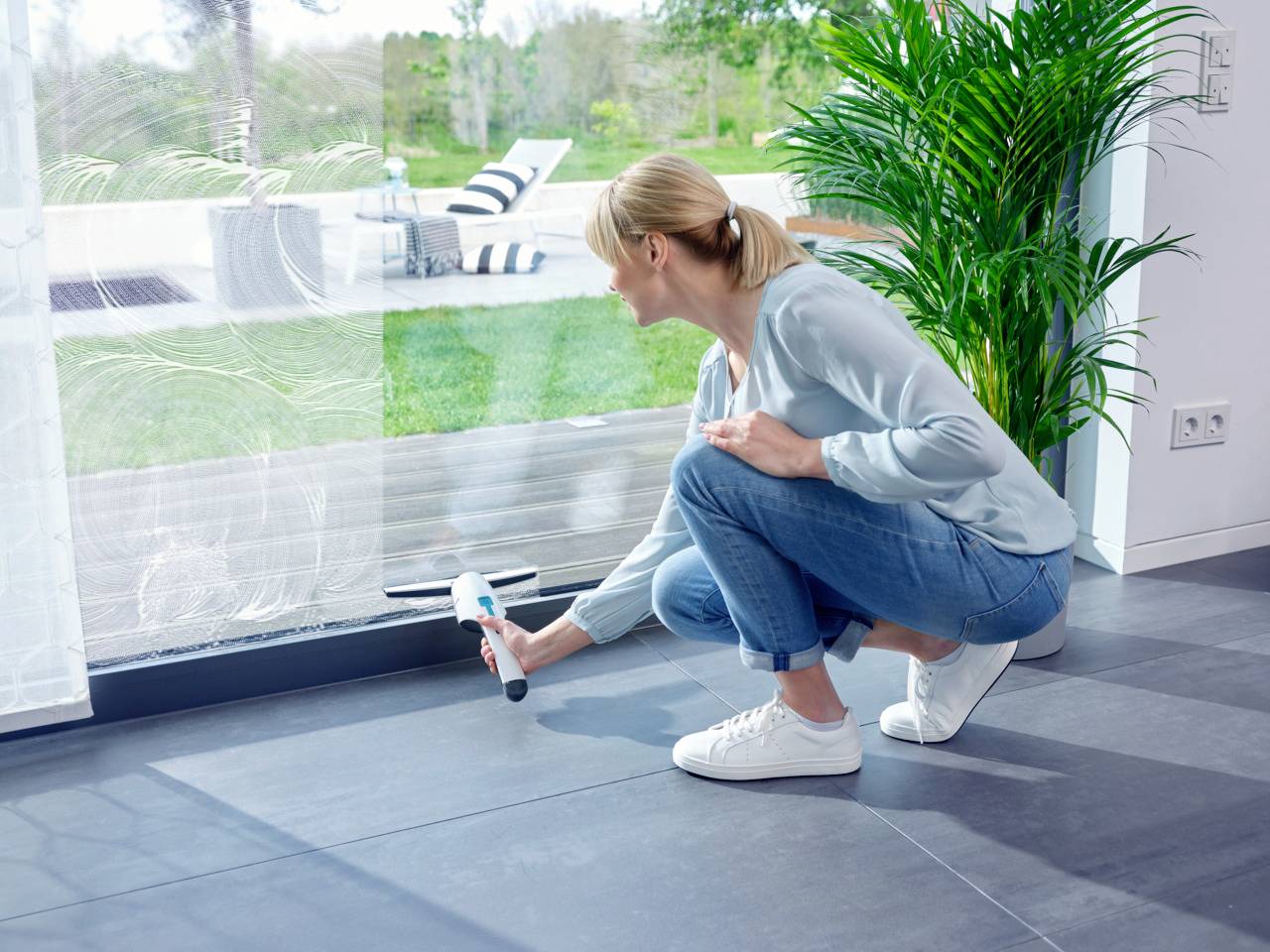 Une femme nettoie une grande fenêtre de l'intérieur à l'aide d'un nettoyeur de vitres électrique. Une terrasse est visible en arrière-plan.