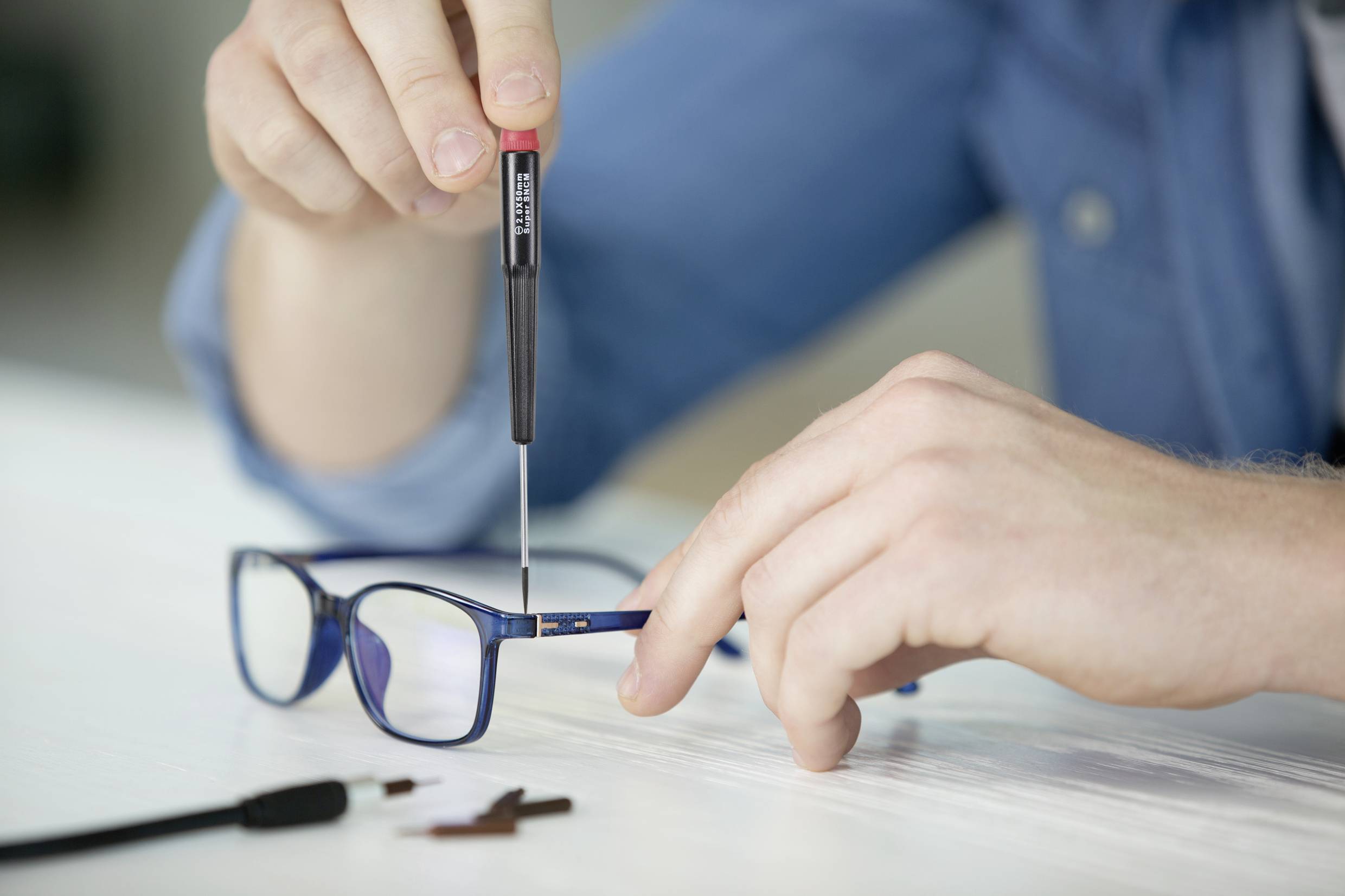 Une personne répare des lunettes avec un petit tournevis. Les lunettes sont posées sur une table, et des pièces de rechange sont à côté.