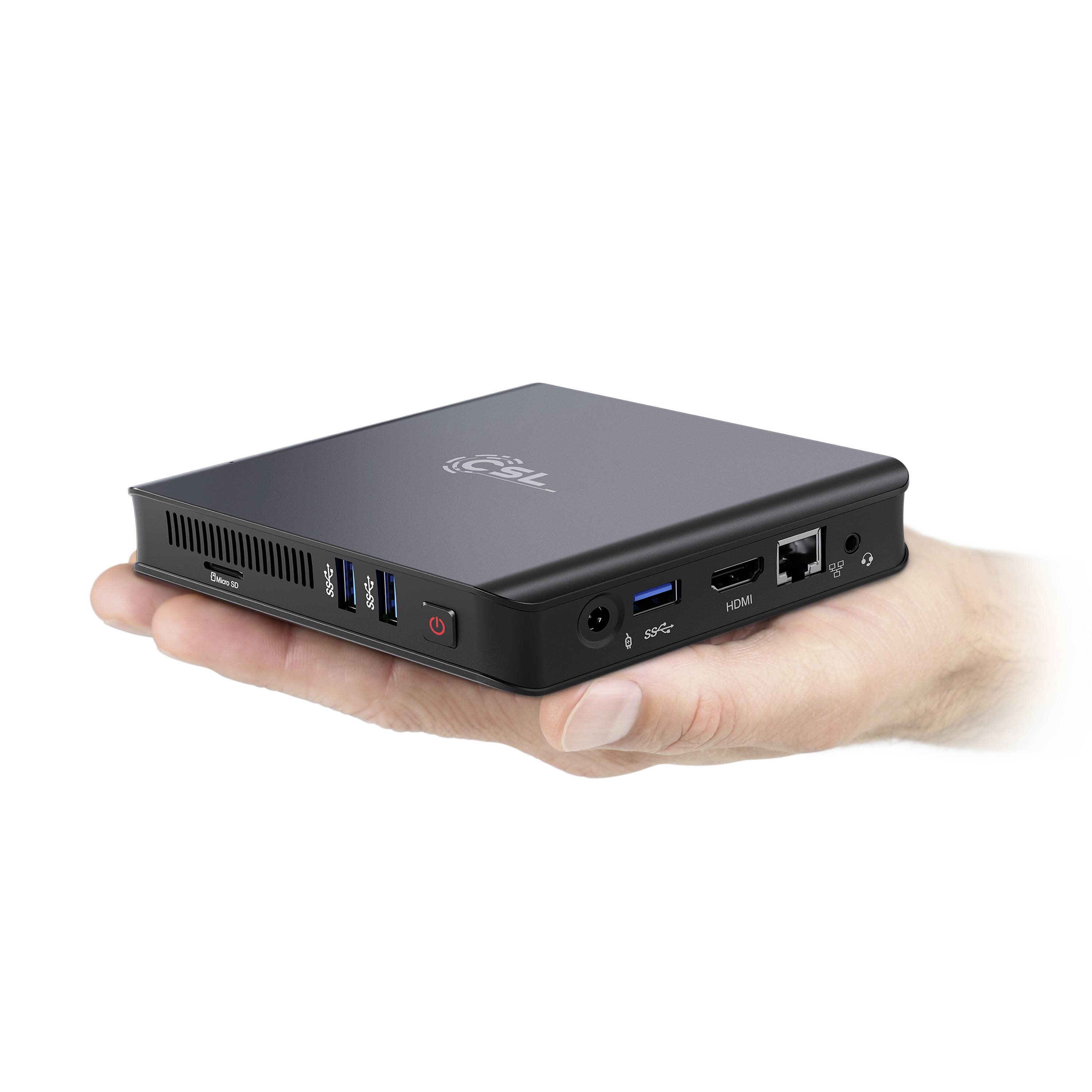 Une main tient un mini-PC compact noir avec plusieurs ports à l'arrière, notamment USB, HDMI et Ethernet.
