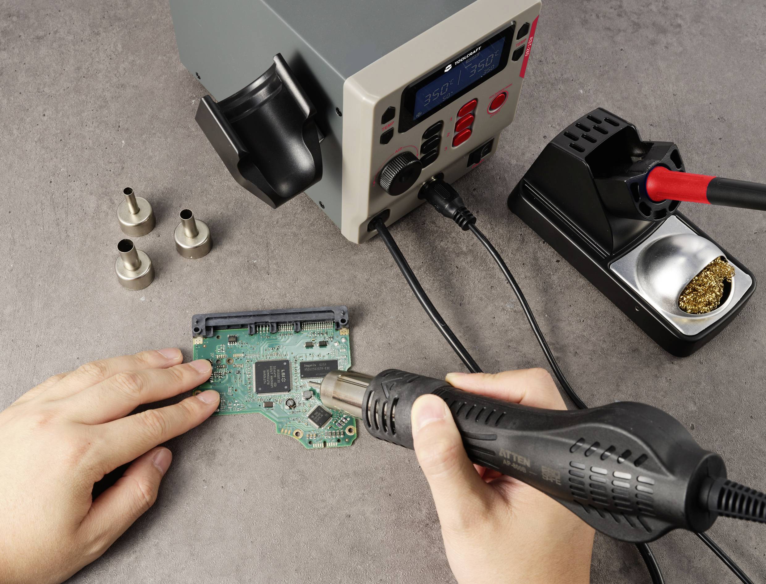 Une personne utilise un pistolet à air chaud pour souder sur une carte de circuit imprimé. Des outils et accessoires sont disposés sur la table à côté de la carte.