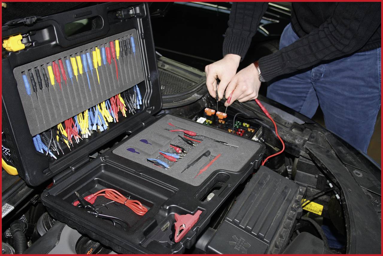 Une personne travaille dans le compartiment moteur ouvert d'une voiture avec des outils de diagnostic sortis d'une mallette à outils. Plusieurs câbles sont visibles.