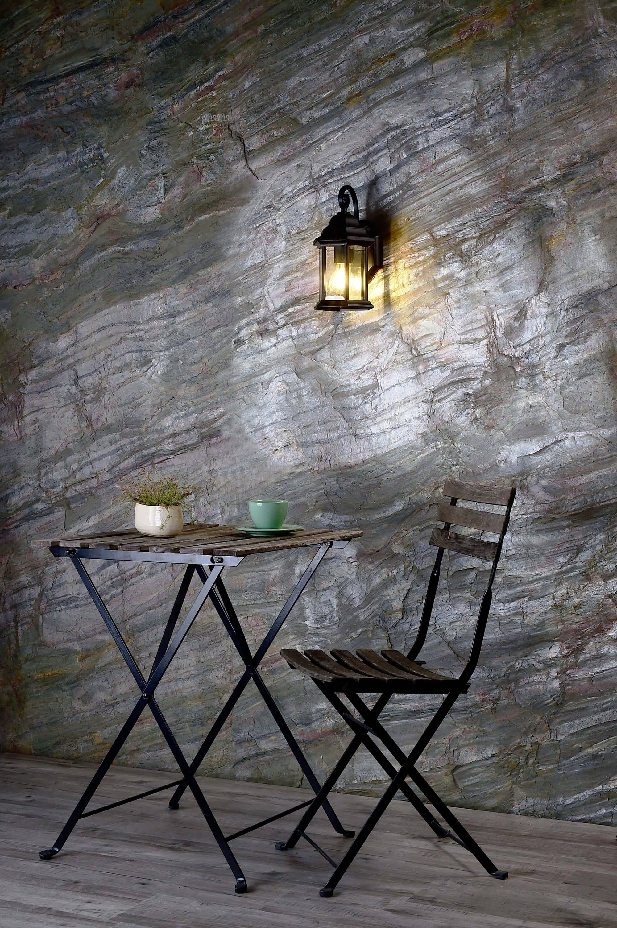 Petite table avec une tasse et une plante à côté, chaise en bois contre un mur de pierre avec une lanterne illuminée.