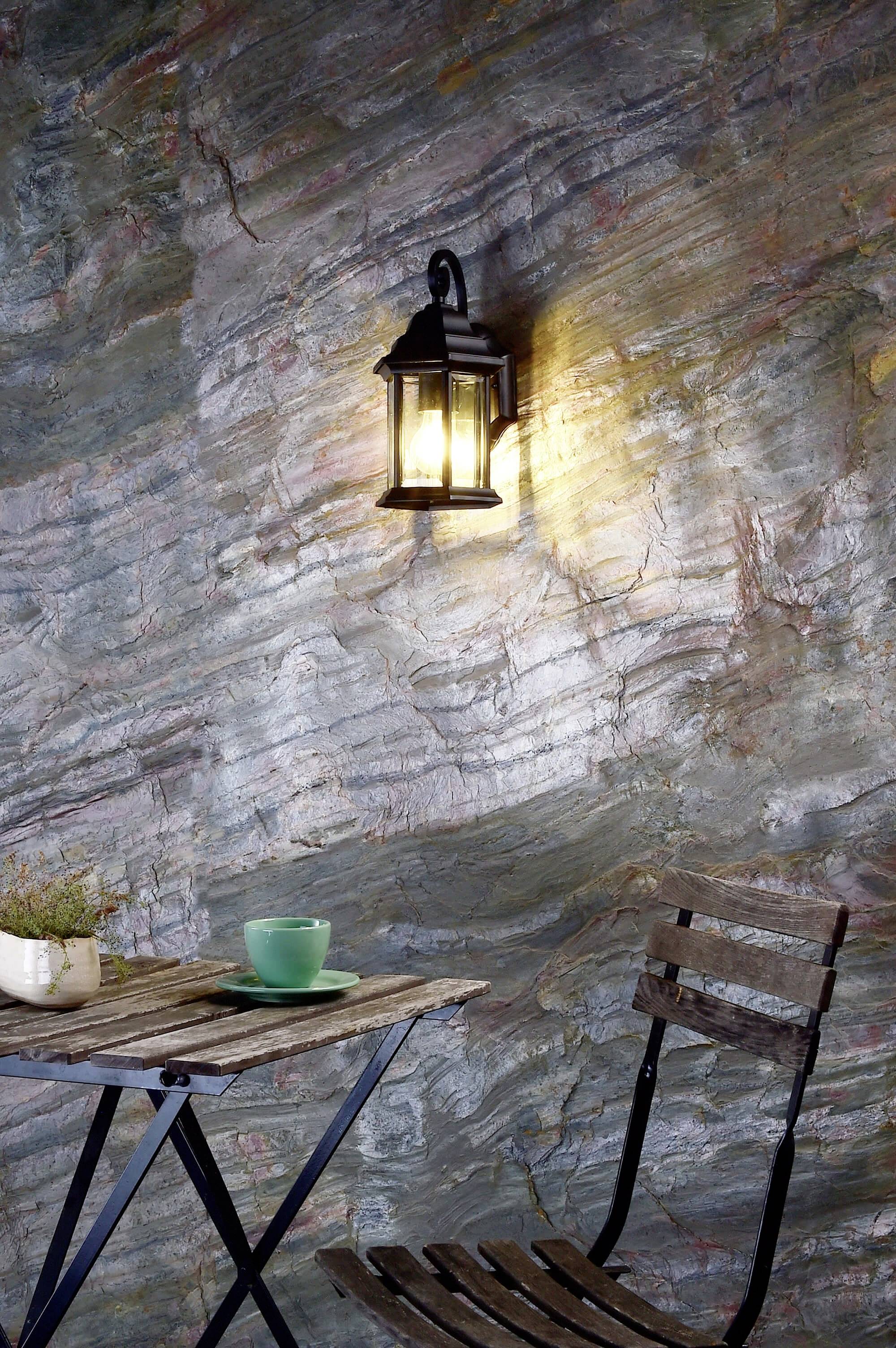 Un support de lampe murale rustique éclaire un mur de pierre. En dessous, une table avec une tasse de thé et une chaise, créant une ambiance chaleureuse et accueillante.