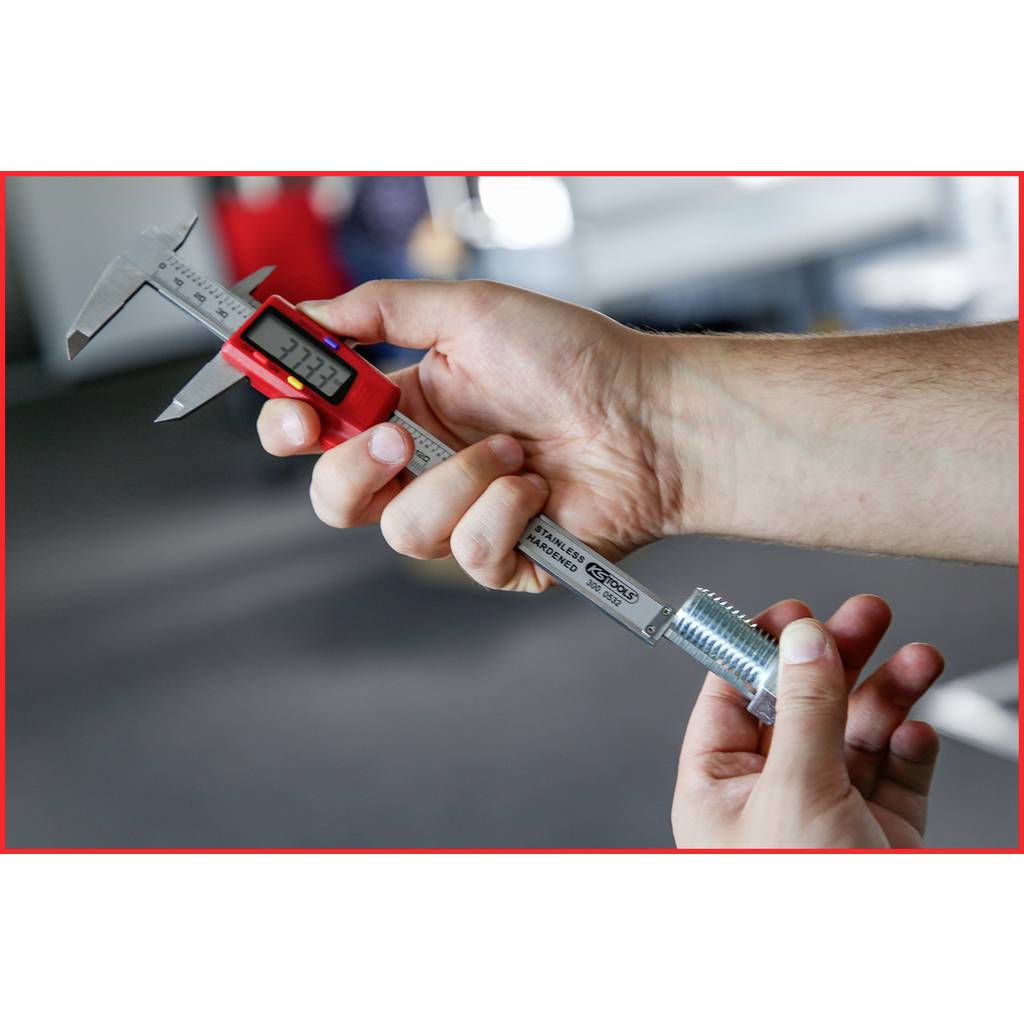 Une personne mesure une pièce métallique à l'aide d'un pied à coulisse numérique. Le pied à coulisse indique 34,22 mm. L'environnement semble industriel.
