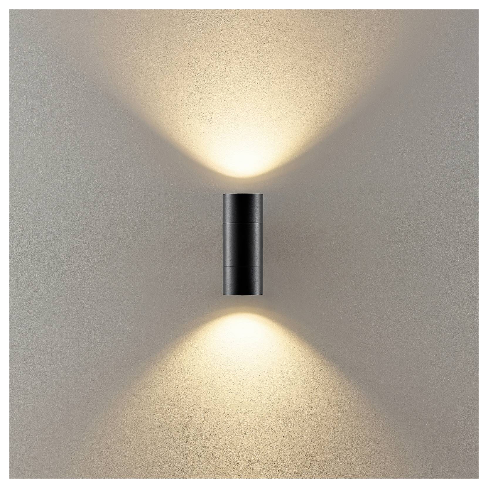 Une applique murale moderne diffuse une lumière chaleureuse vers le haut et vers le bas sur un mur beige.
