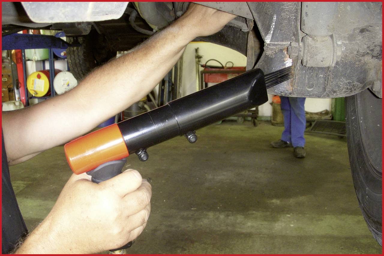Une personne utilise un outil pneumatique pour enlever la rouille d'un châssis de véhicule dans un atelier. Des outils sont visibles en arrière-plan.