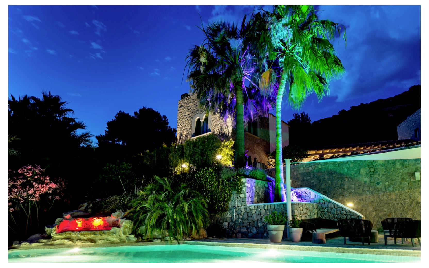 Scène de station tropicale nocturne avec palmiers illuminés, bâtiment en pierre, piscine et éclairage paysager coloré sous un ciel bleu profond.