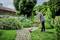 Un homme tond la pelouse avec un coupe-bordures électrique sur un chemin de jardin bien entretenu, entouré de feuillage vert et d'une maison en bois en arrière-plan.