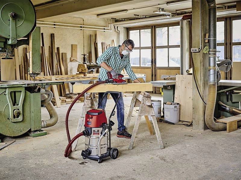 Une personne travaille dans un atelier de menuiserie avec un rabot électrique pour lisser une planche de bois. Des machines et des stocks de bois sont visibles en arrière-plan.