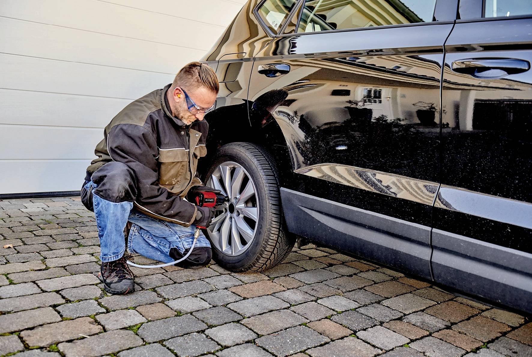 Un homme est à genoux sur une allée pavée et change le pneu d'une voiture noire à l'aide d'une visseuse électrique.
