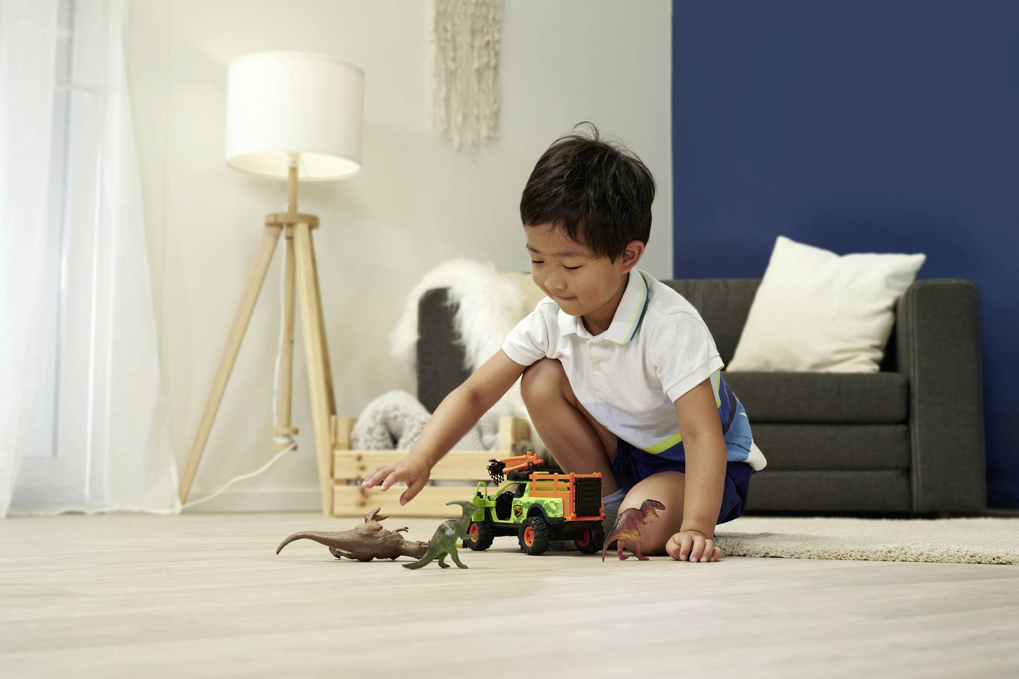 Un enfant joue sur le sol d'un salon avec des jouets de dinosaures et un petit tracteur. Un canapé et une lampe sont visibles en arrière-plan.