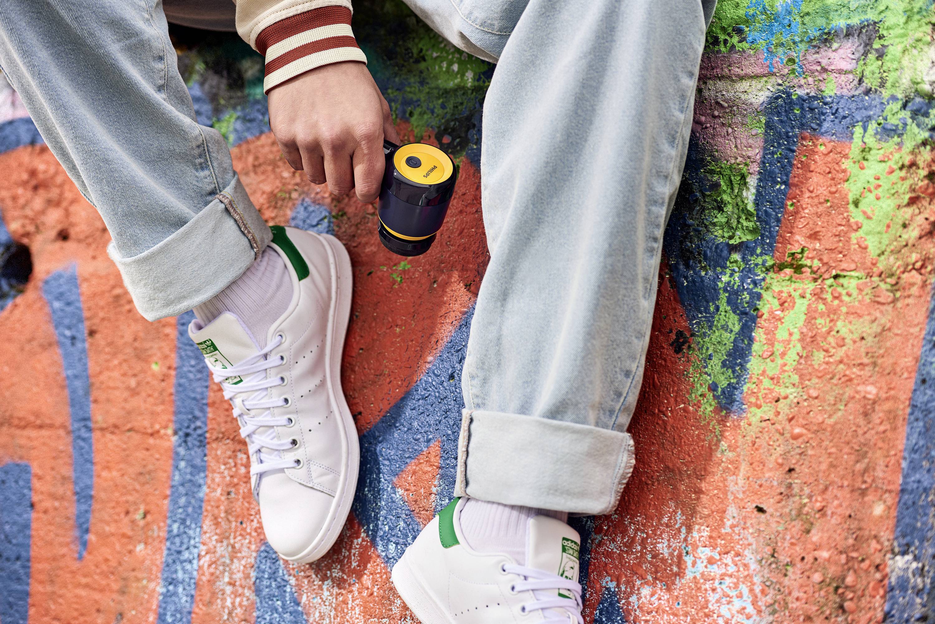Une personne est assise nonchalamment sur un mur coloré, recouvert de graffitis, et tient une petite enceinte. Elle porte un jean et des baskets blanches.