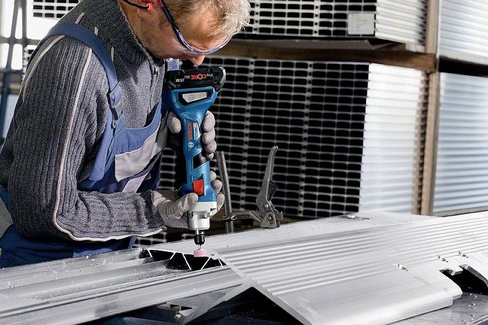 Une personne travaille une grande tôle métallique avec un outil électrique dans un environnement d'atelier. Elle porte des lunettes de protection et des vêtements de travail.