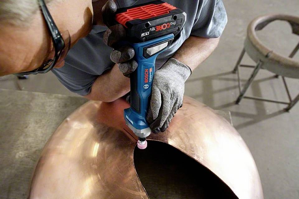 Une personne utilise un outil de ponçage électrique pour polir la surface d'un grand objet de couleur cuivrée.