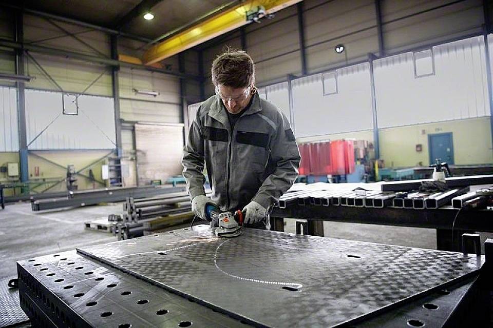 Une personne ponçe une grande plaque métallique dans un hall d'usine. Elle porte des vêtements de protection et travaille avec concentration sur une table.