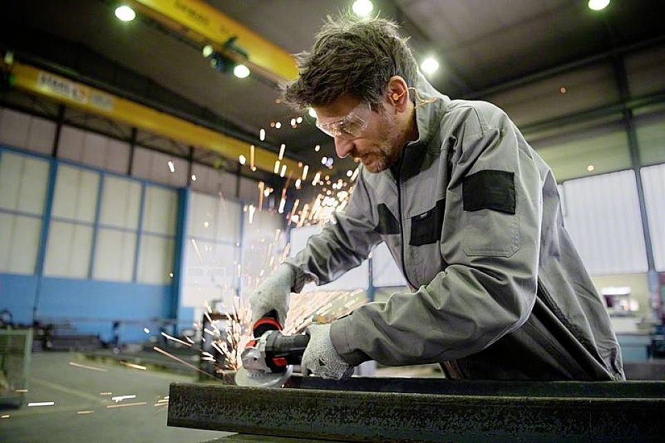 Un homme meule une pièce métallique avec une meuleuse d'angle dans un atelier. Des étincelles jaillissent. Il porte des lunettes de protection et des vêtements de travail.