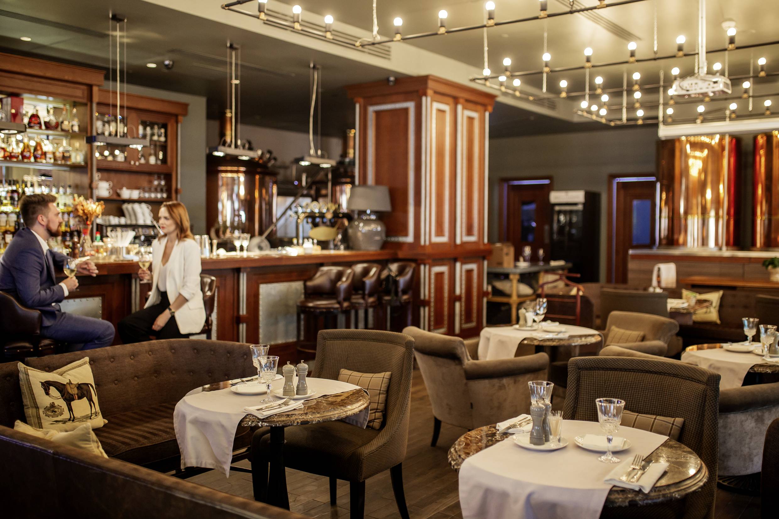 Un bar élégant avec une décoration en bois et des lampes modernes. À l'avant, des tables confortables sont installées, et à l'arrière, un couple discute au comptoir.