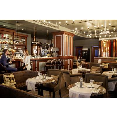 Un bar élégant avec une décoration en bois et des lampes modernes. À l'avant, des tables confortables sont installées, et à l'arrière, un couple discute au comptoir.