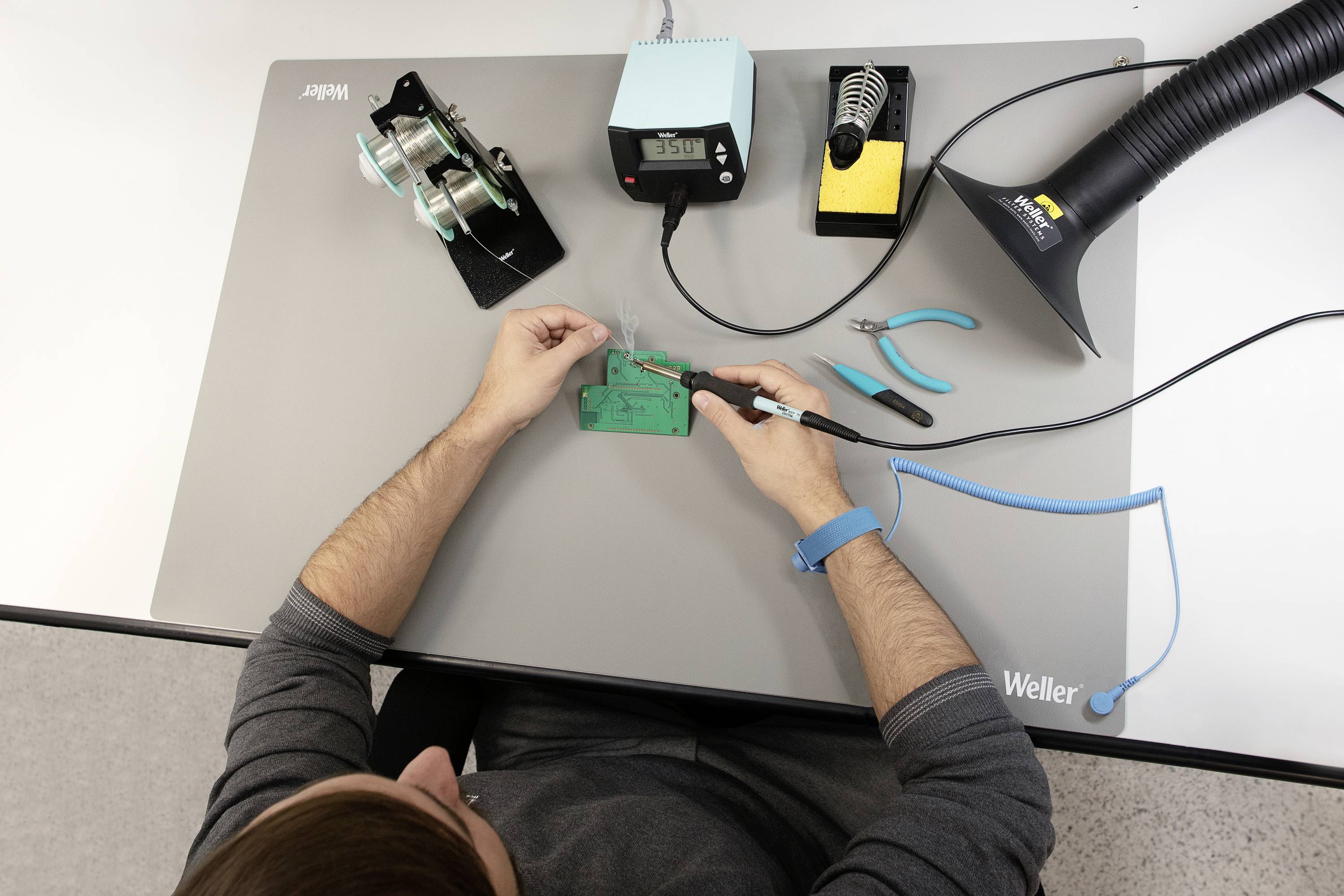 Une personne soude une carte électronique verte sur une table, équipée d'outils de soudure et d'un système d'aspiration. Sur la table se trouvent des bobines de fil et des pinces.