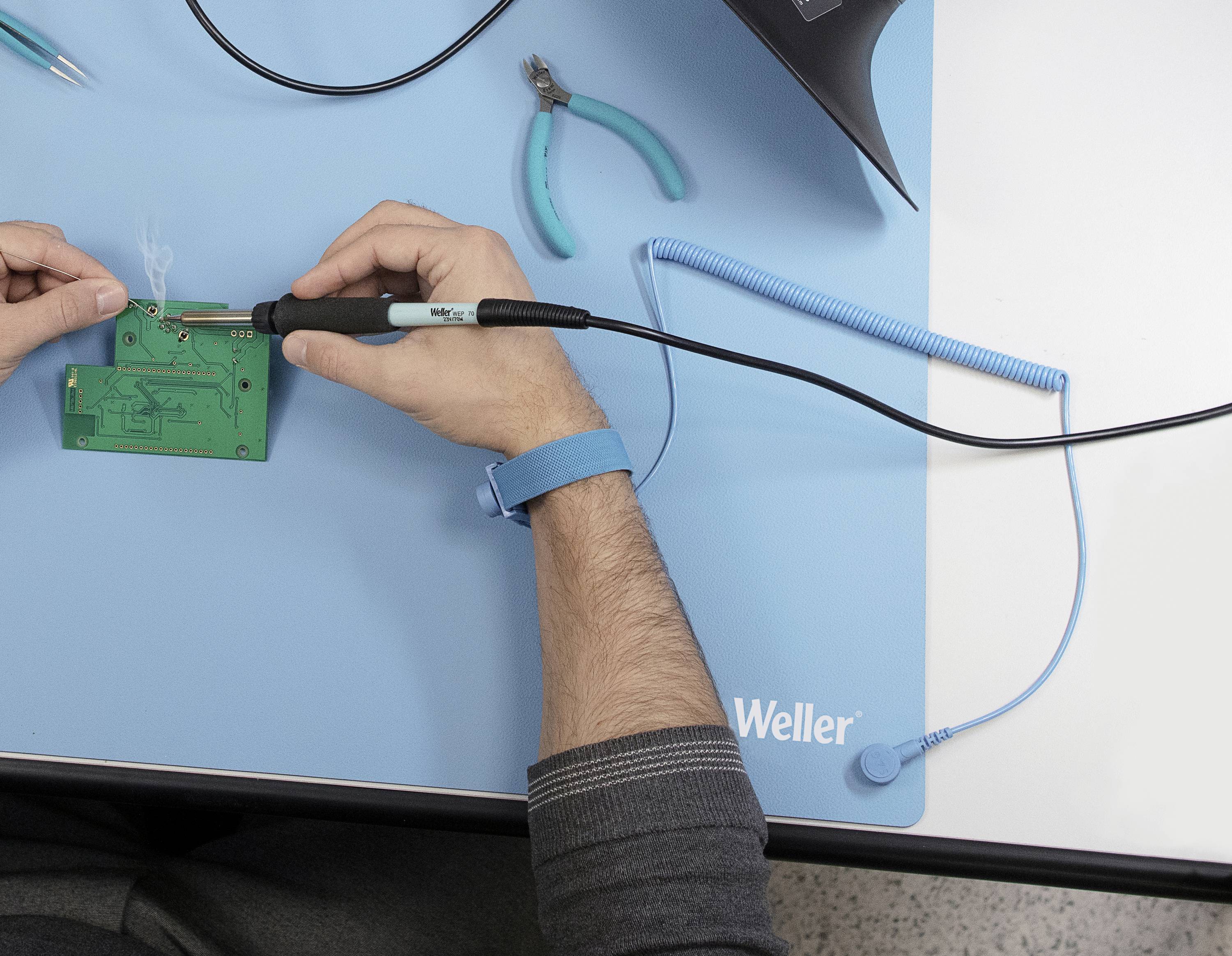 Une personne soude un circuit imprimé vert sur un tapis bleu à l'aide d'un fer à souder. Des outils sont visibles en arrière-plan.