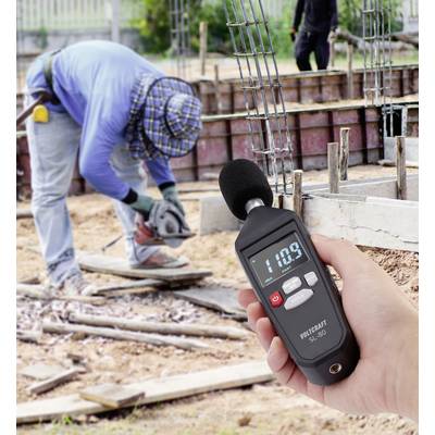 Un appareil de mesure des décibels affiche '110,9', alors qu'en arrière-plan, une personne travaille avec une scie sur un chantier de construction.