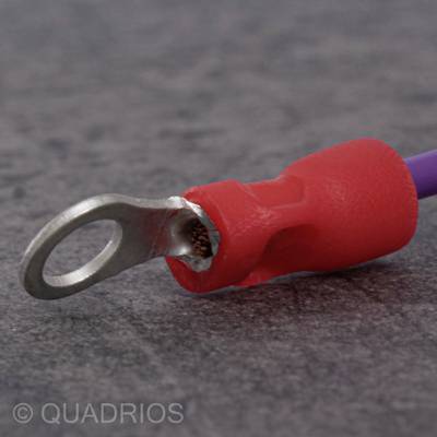 Un cosses de câble rouge avec un œillet de connexion est posé sur une surface grise. Il est utilisé pour connecter des conducteurs électriques.