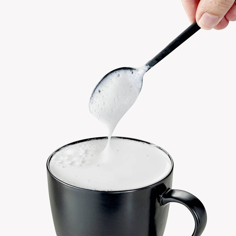Une main tient une cuillère pleine de mousse de lait au-dessus d'une tasse noire remplie de mousse de lait.
