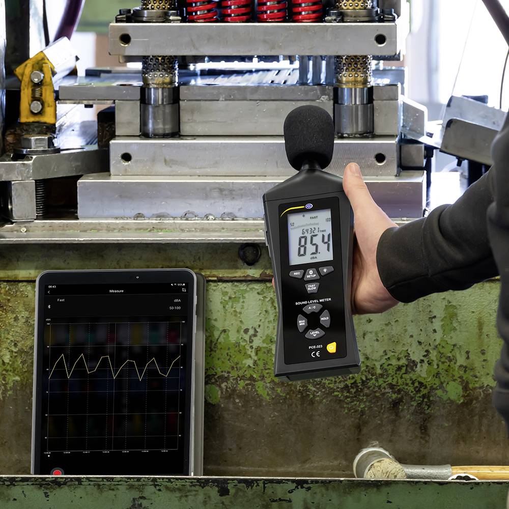 Un appareil portable mesure les valeurs en décibels devant une machine, affichant 83,4. Une tablette à côté montre une courbe du niveau sonore.